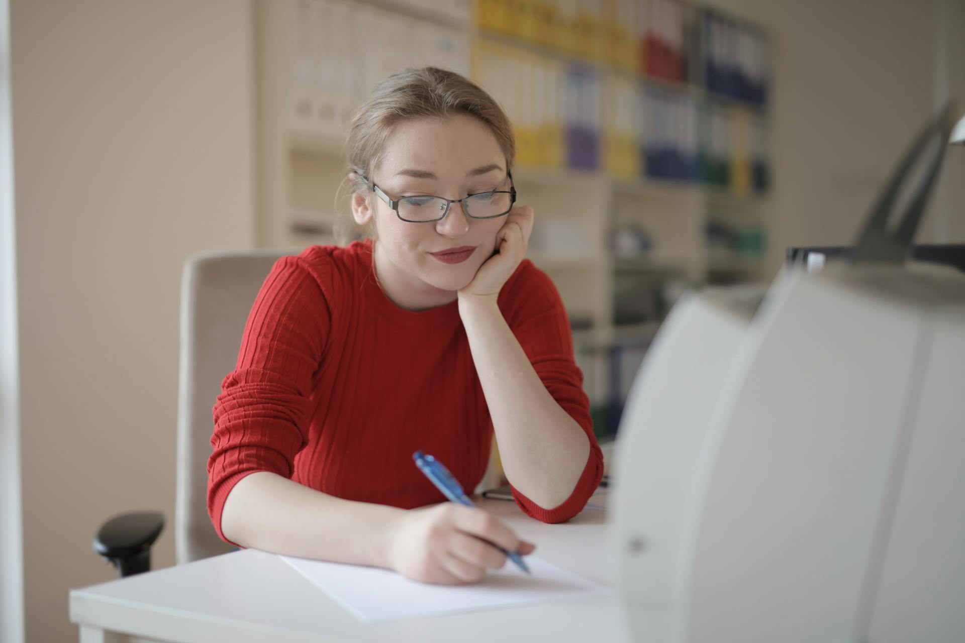 A woman in glasses is writing at a desk in an office setting.