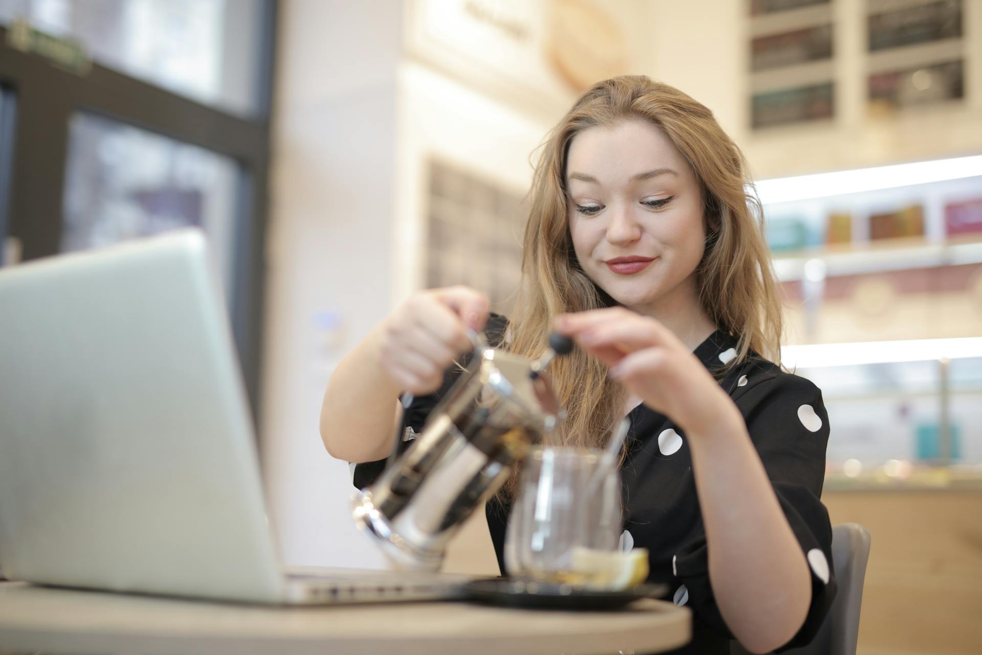 Relaxed female in trendy polka dot dress pouring hot beverage from french press into glass mug while chilling in cafe during weekend