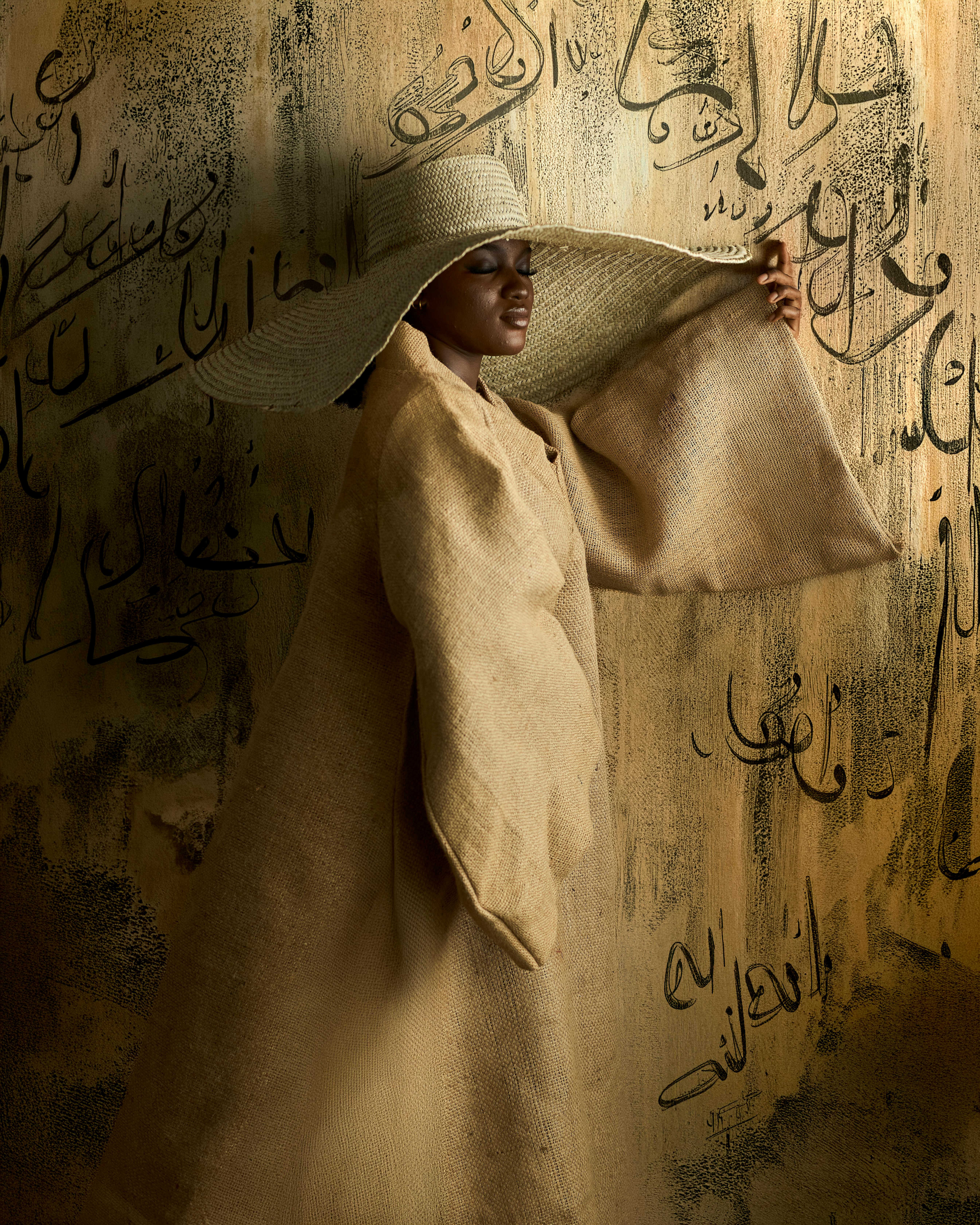 Stylish woman in a wide-brimmed hat and beige cloak poses against calligraphy wall.