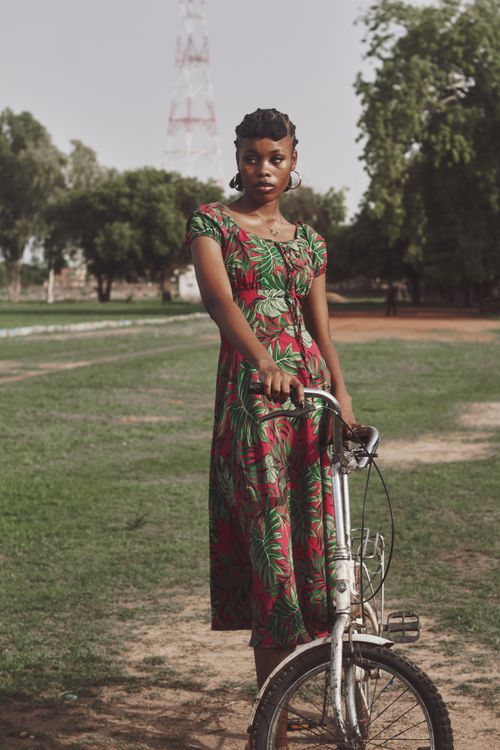 Free A young woman in a colorful floral dress stands with her bicycle on a sunny day in Zaria, Nigeria. Stock Photo