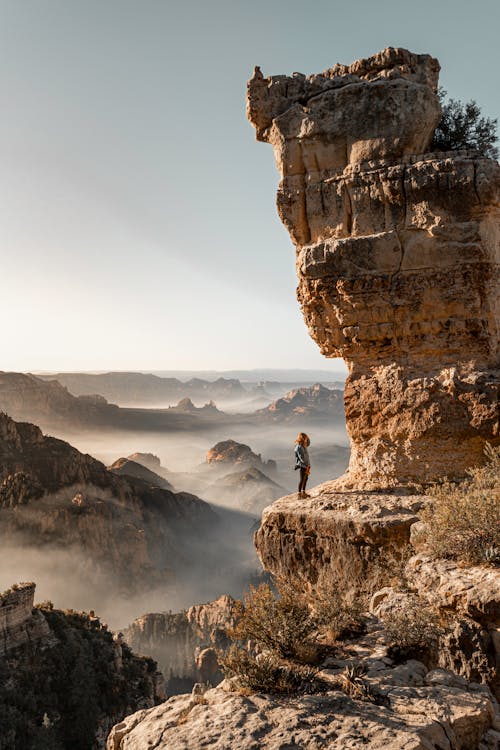 Side view of anonymous female traveler standing on shabby mount under high shapeless cliff under serene sky on foggy weather during trip