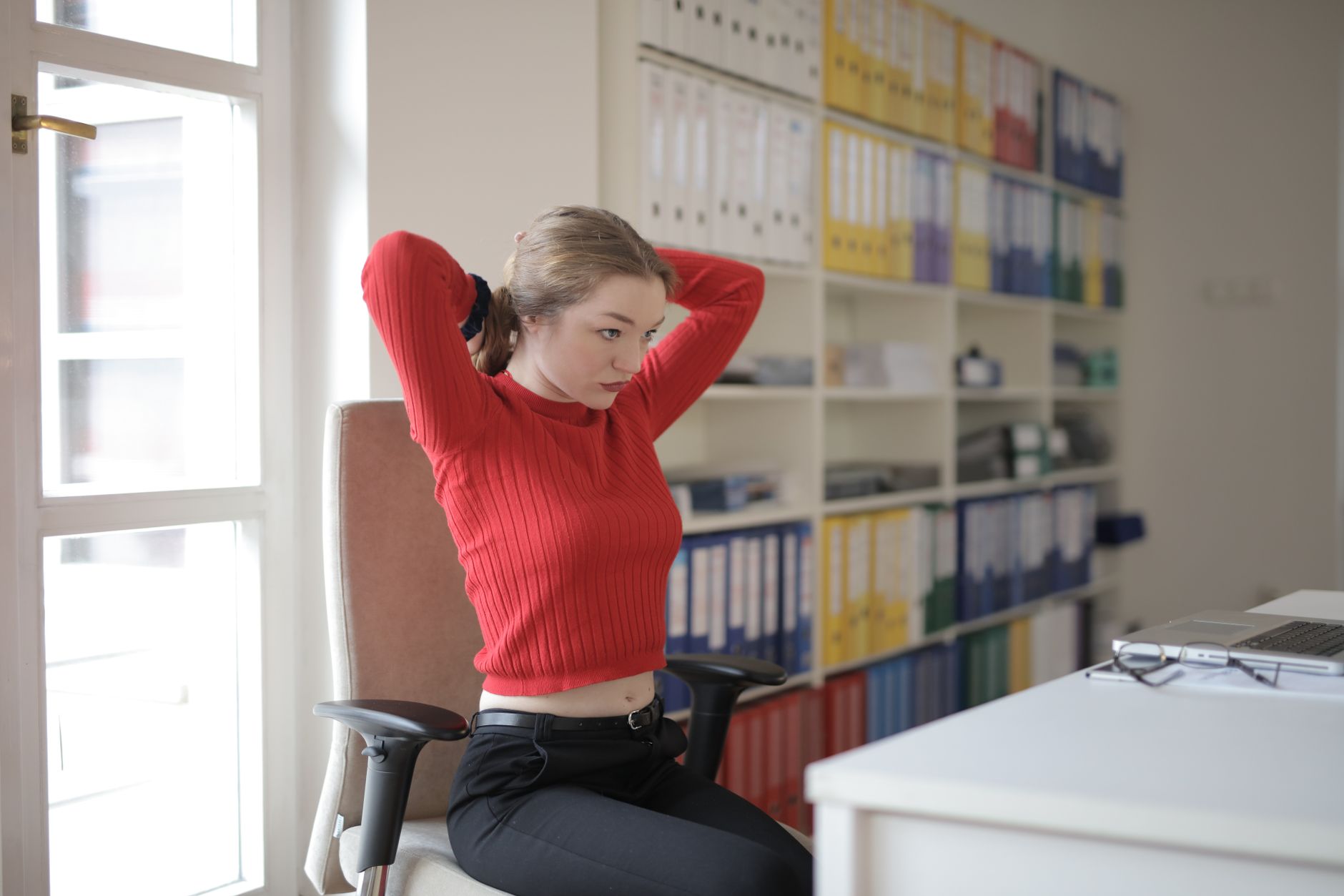 Emotionless female secretary in casual wear sitting at table in contemporary workplace and tying ponytail during work