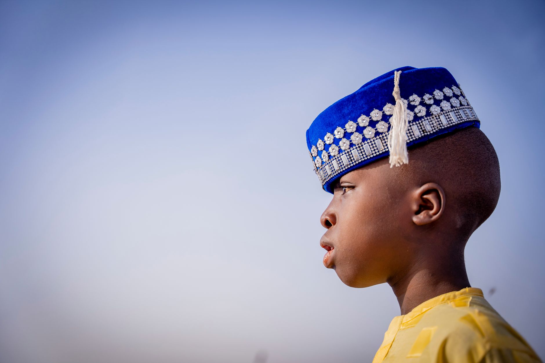 Close-up of a young boy wearing a traditional blue cap, gazing outdoors under a clear sky in Kano, Nigeria.