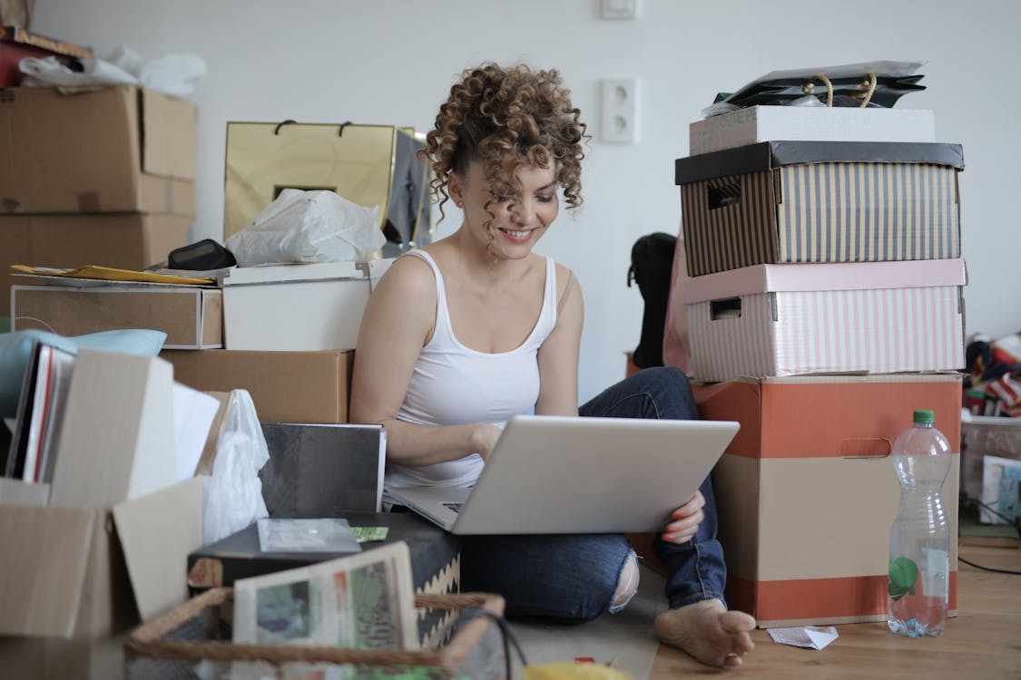 Free Smiling female customer in casual wear sitting on floor in untidy room with stacks of cardboard boxes and making orders in online store while using laptop Stock Photo
