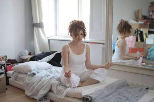 Tranquil female in sleepwear sitting on bed in messy room and practicing yoga with closed eyes while maintaining mental health