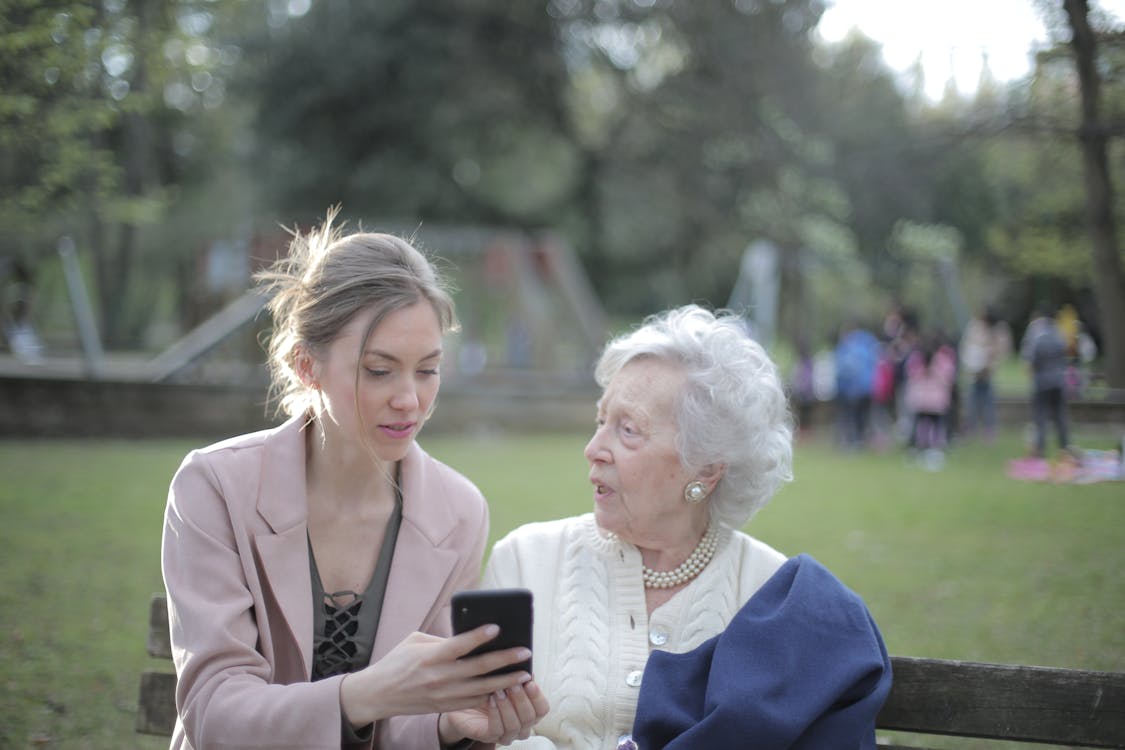 Free Adult daughter teaching senior mother using smartphone in park Stock Photo