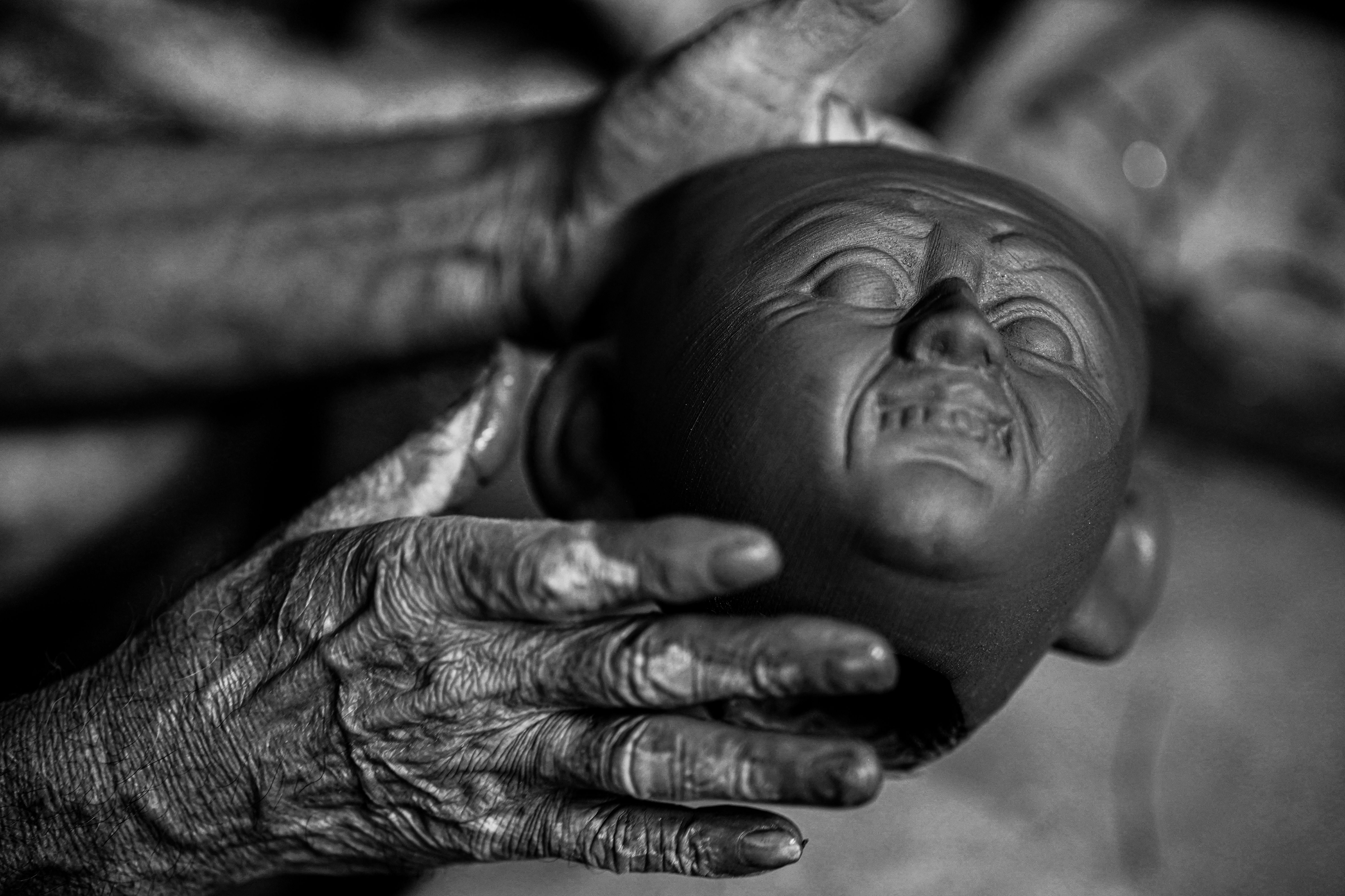 A close-up of an elderly artisan's hands crafting a clay sculpture head, in a traditional setting in Bangladesh.