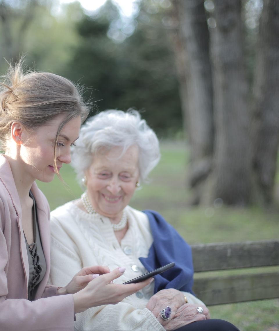 A patient caregiver sharing a warm, reassuring smile with an older woman in a sunlit living space.