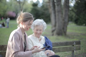 Side view of smiling adult female helping aged mom in using of mobile phone while sitting together in park