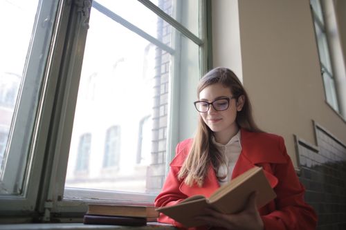 Free A young woman in a red coat happily reading a book by a window indoors. Stock Photo