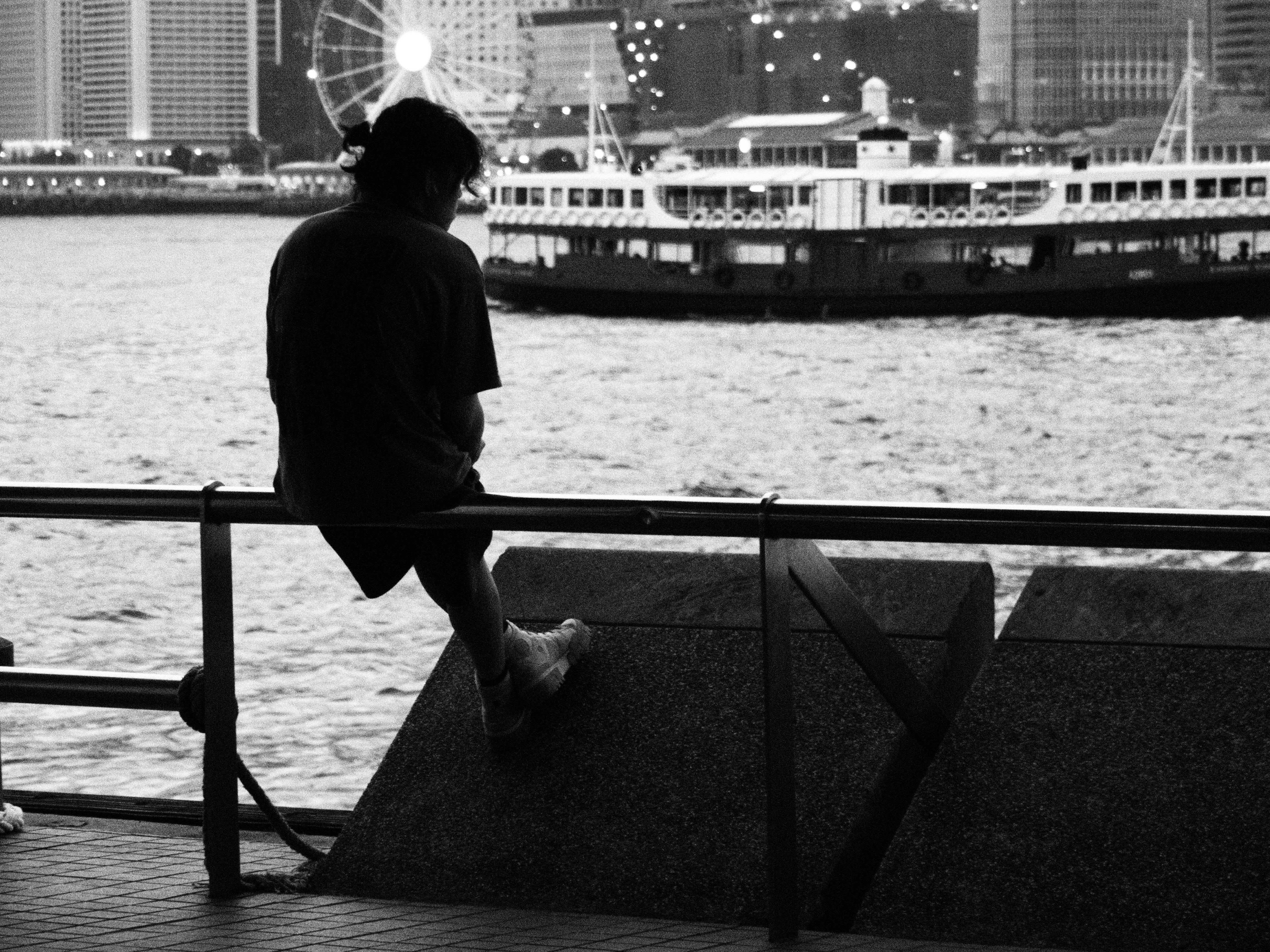 Silhouette of a person sitting by Hong Kong Harbor with a ferry and cityscape in the background.