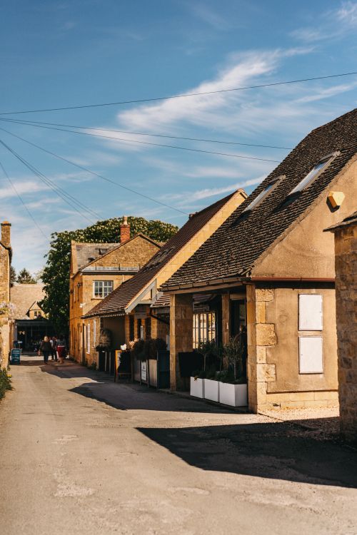 Free Charming village street scene in Cotswolds, England with traditional stone houses under a bright blue sky. Stock Photo