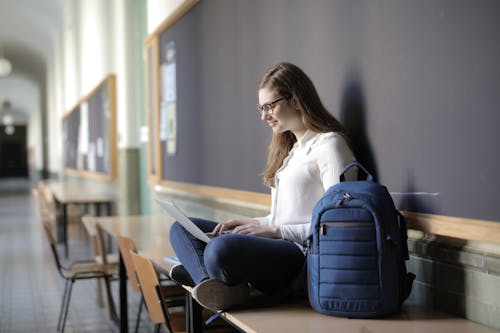 Woman Using Laptop While Sitting on Table Near Blue Backpack