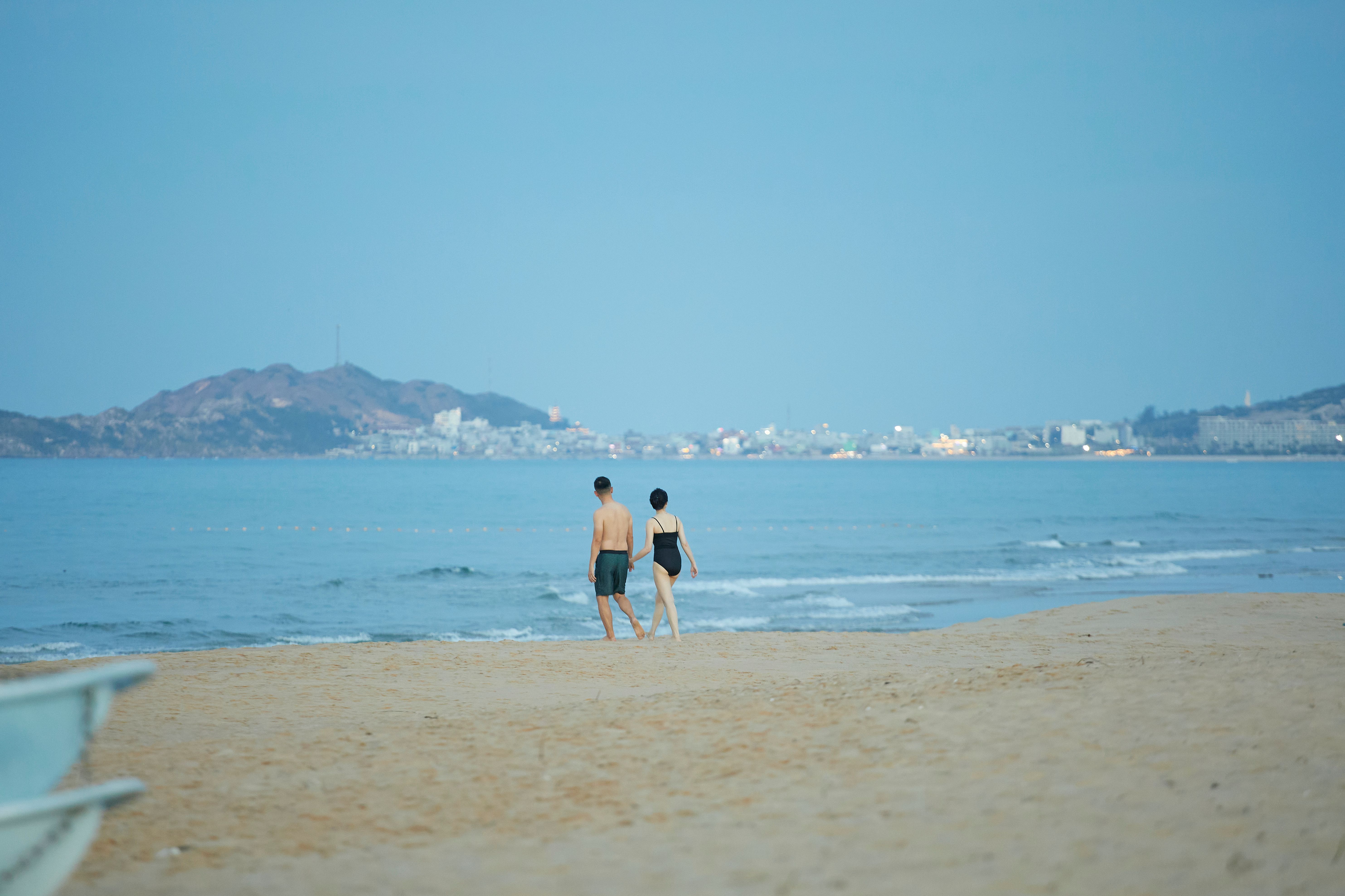 A couple walking along Quy Nhon beach with a distant cityscape under a clear sky.