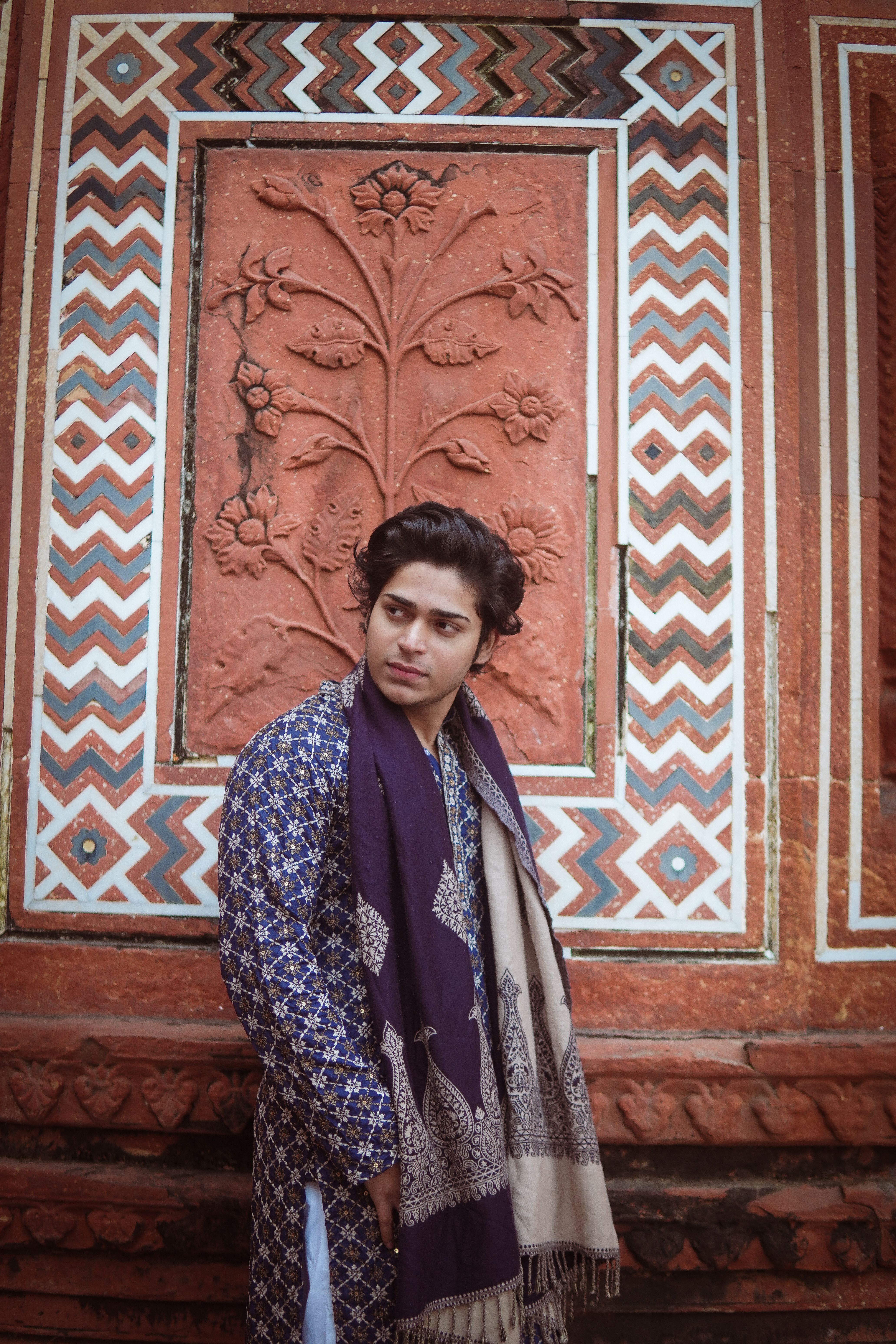 A young man in traditional Indian attire poses against ornate Agra wall.