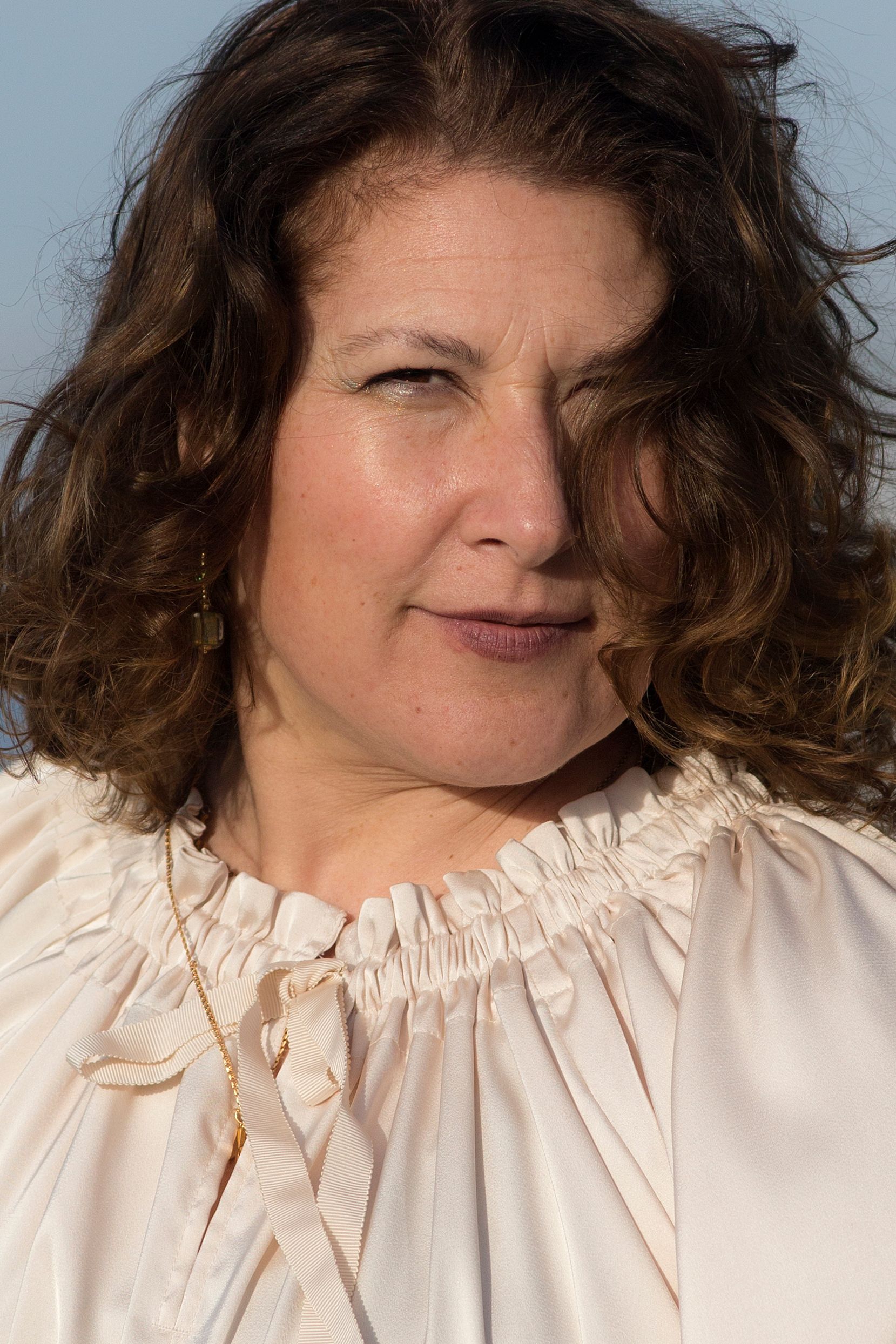 Close-up of a woman with curly hair wearing a white blouse, sunny outdoors.