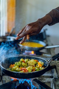 A cook seasons mixed vegetables in a steaming skillet on a stove.