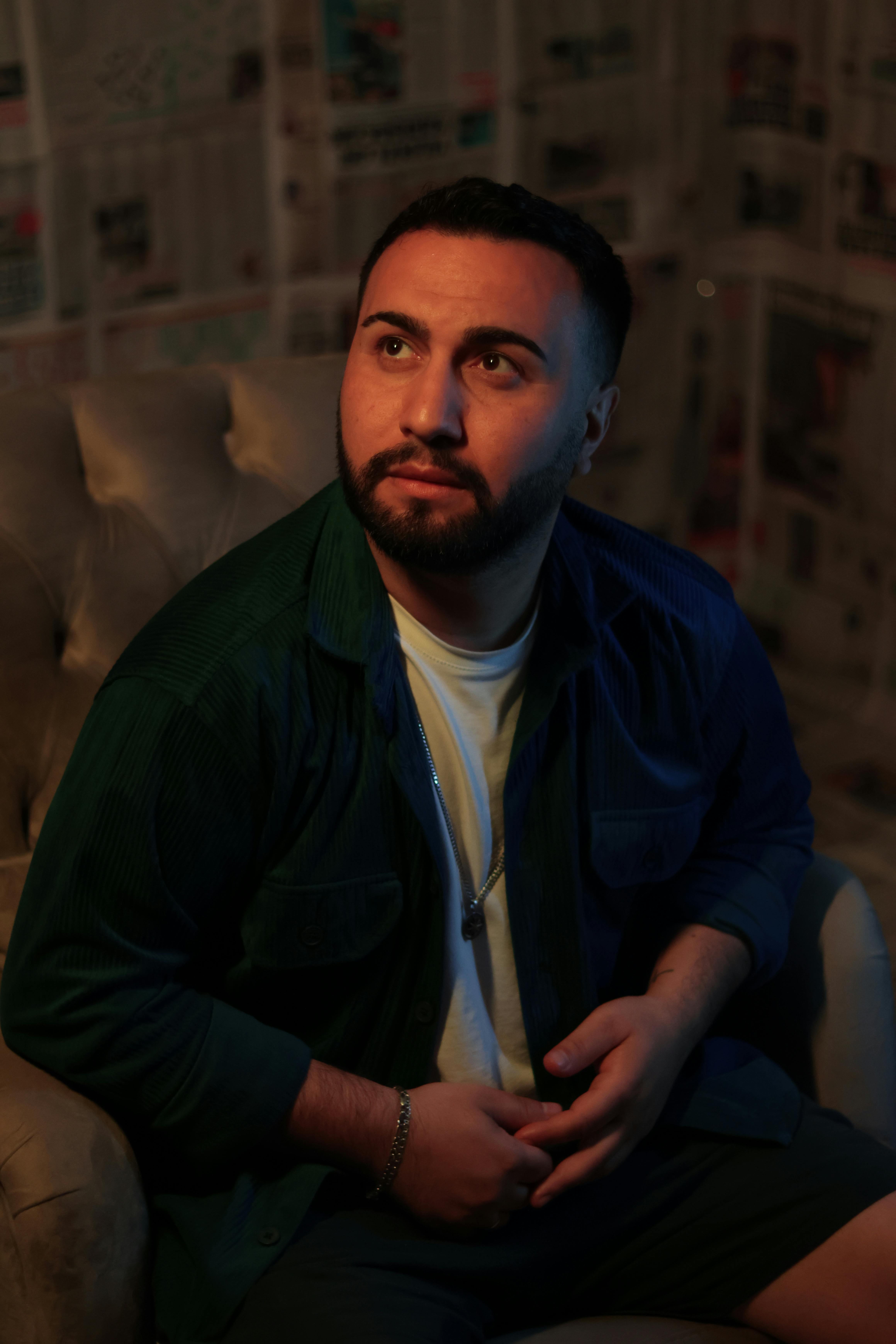 Pensive man seated in a dimly lit room with newspaper walls and soft shadows.