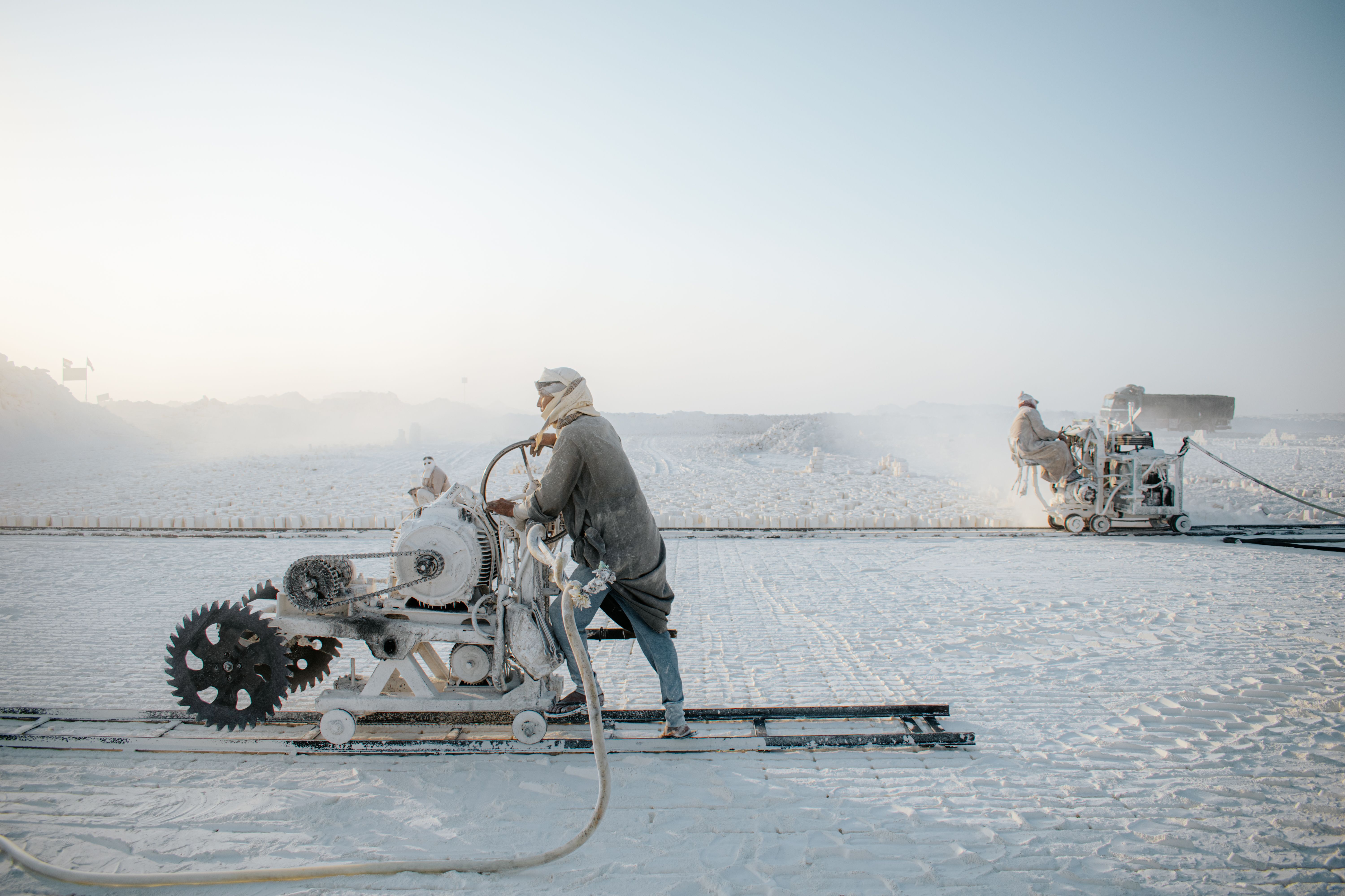 Industrial workers operating equipment in a large open quarry with a white, dusty landscape.