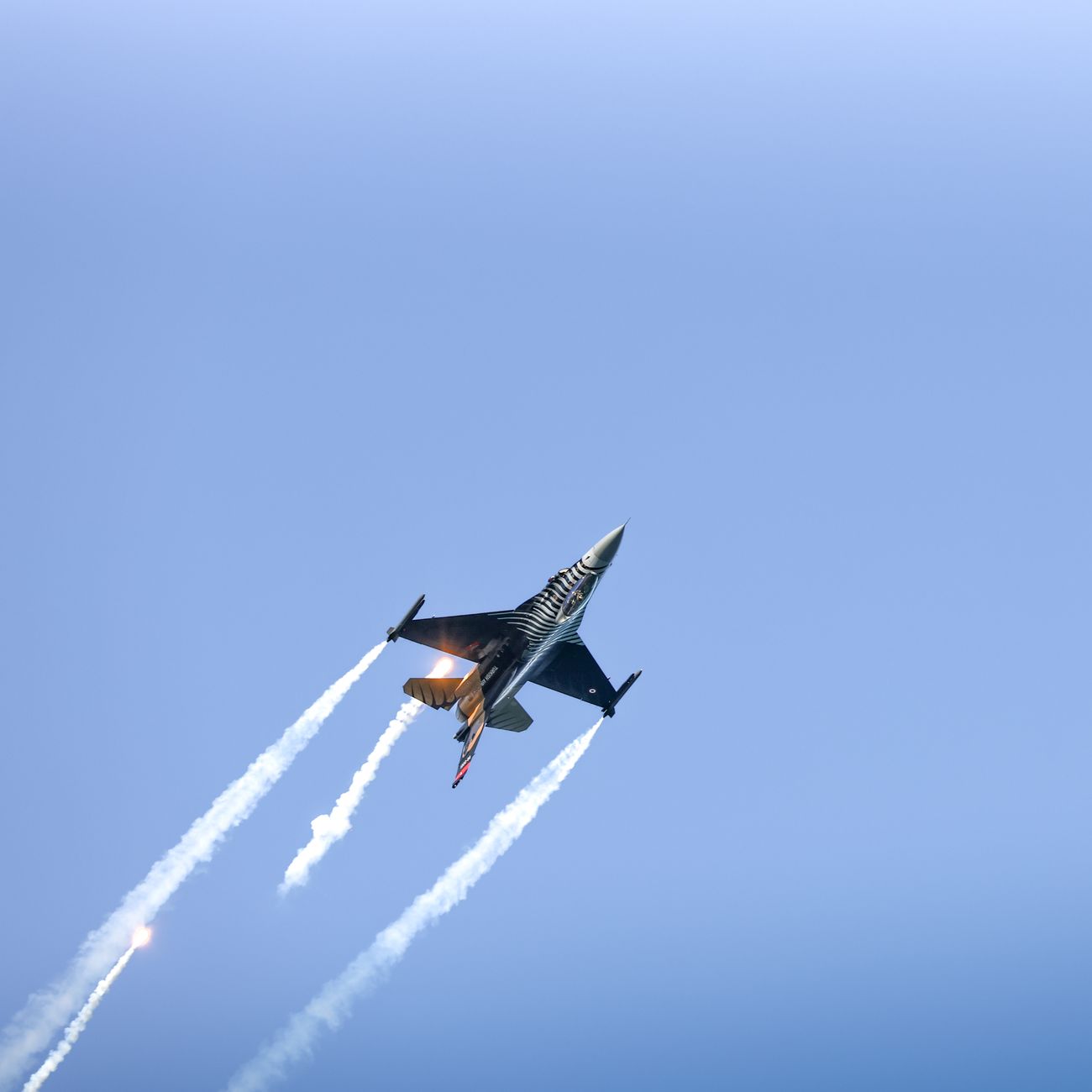 A jet fighter executes an aerial maneuver, releasing smoke trails against a clear blue sky.