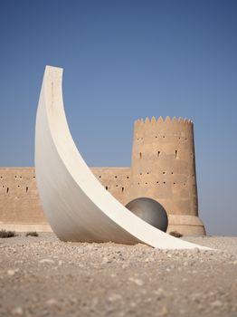 Modern sculpture and historic Al Zubarah Fort, Qatar under a clear blue sky.