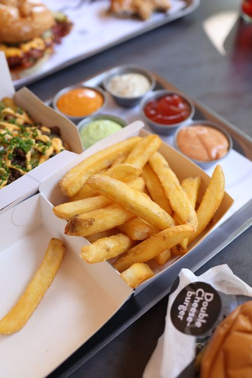 Free Close-up of crispy French fries with a variety of dipping sauces and a burger in the background. Stock Photo