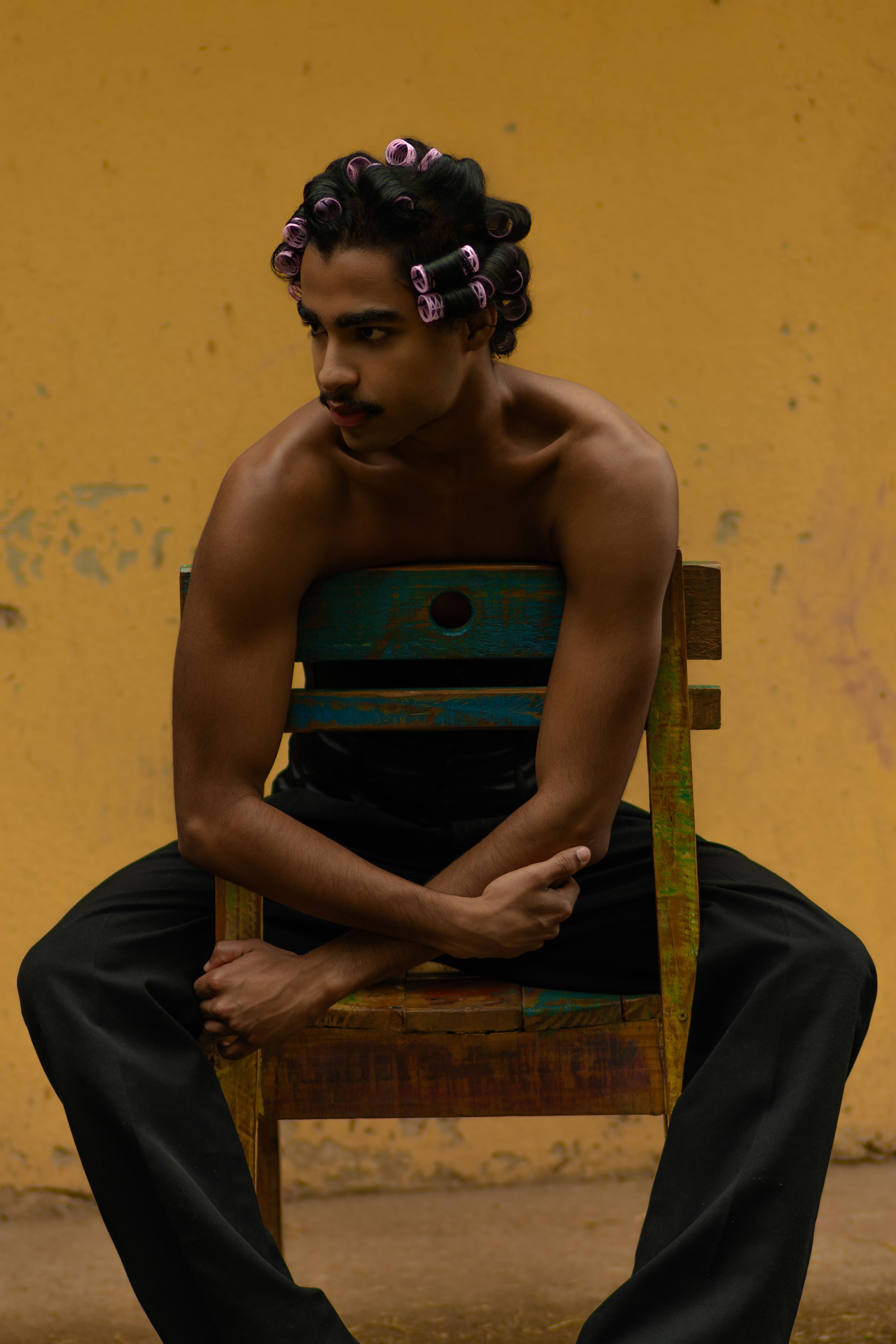 Young man with hair rollers sits on a chair against a warm yellow background, exuding confidence and style.