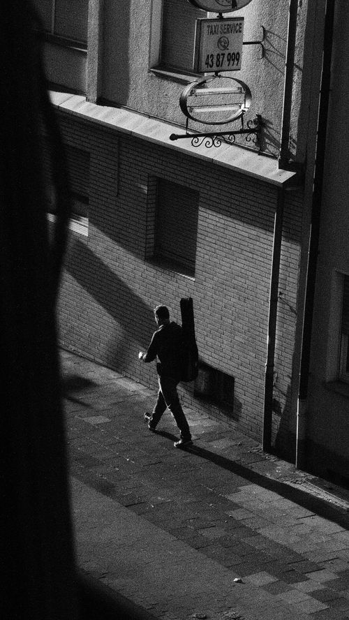 Free Black and white photo of a man walking on a sunlit street with guitar. Stock Photo