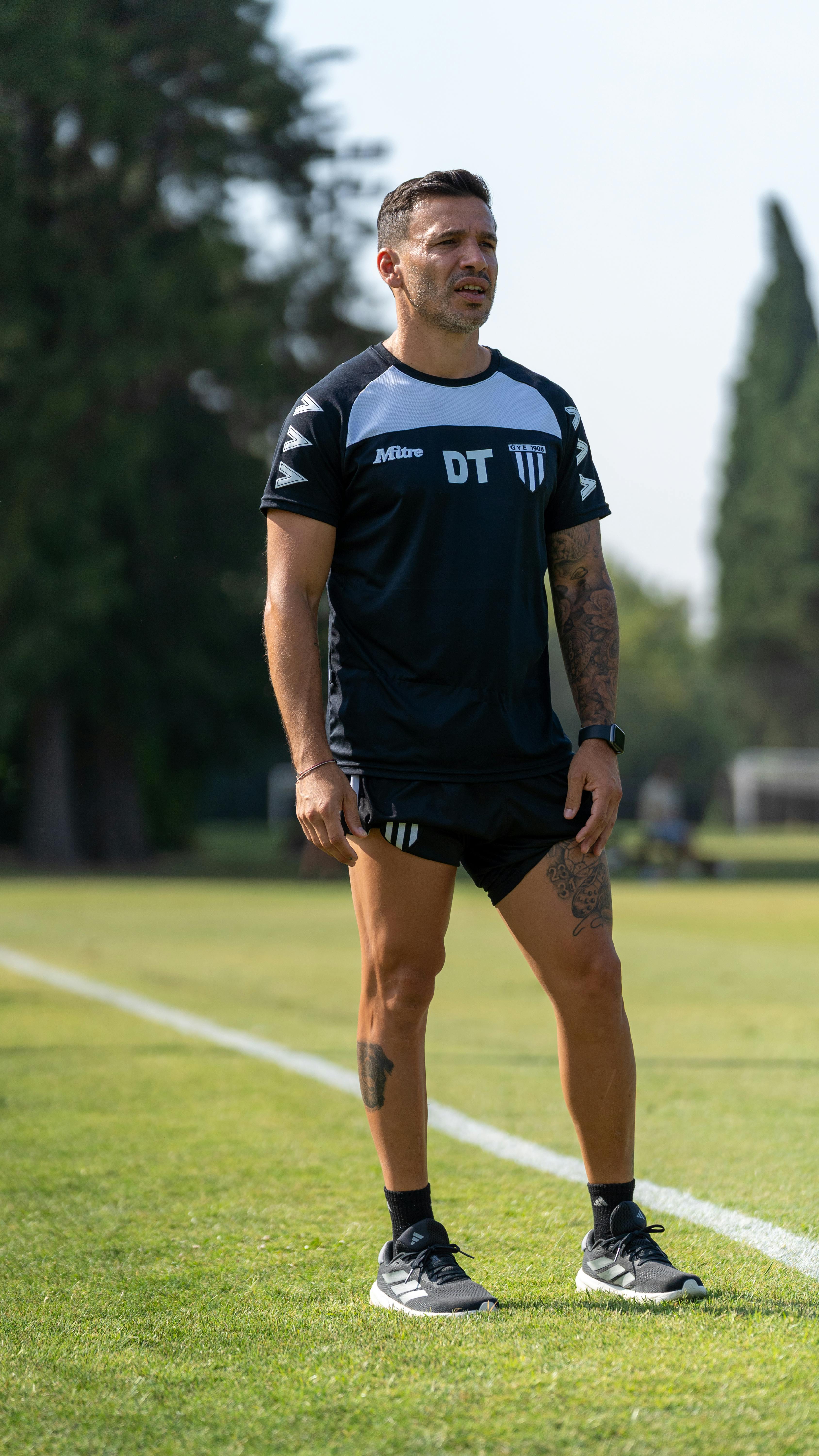 Male soccer coach in casual attire standing on a grass field during day practice.