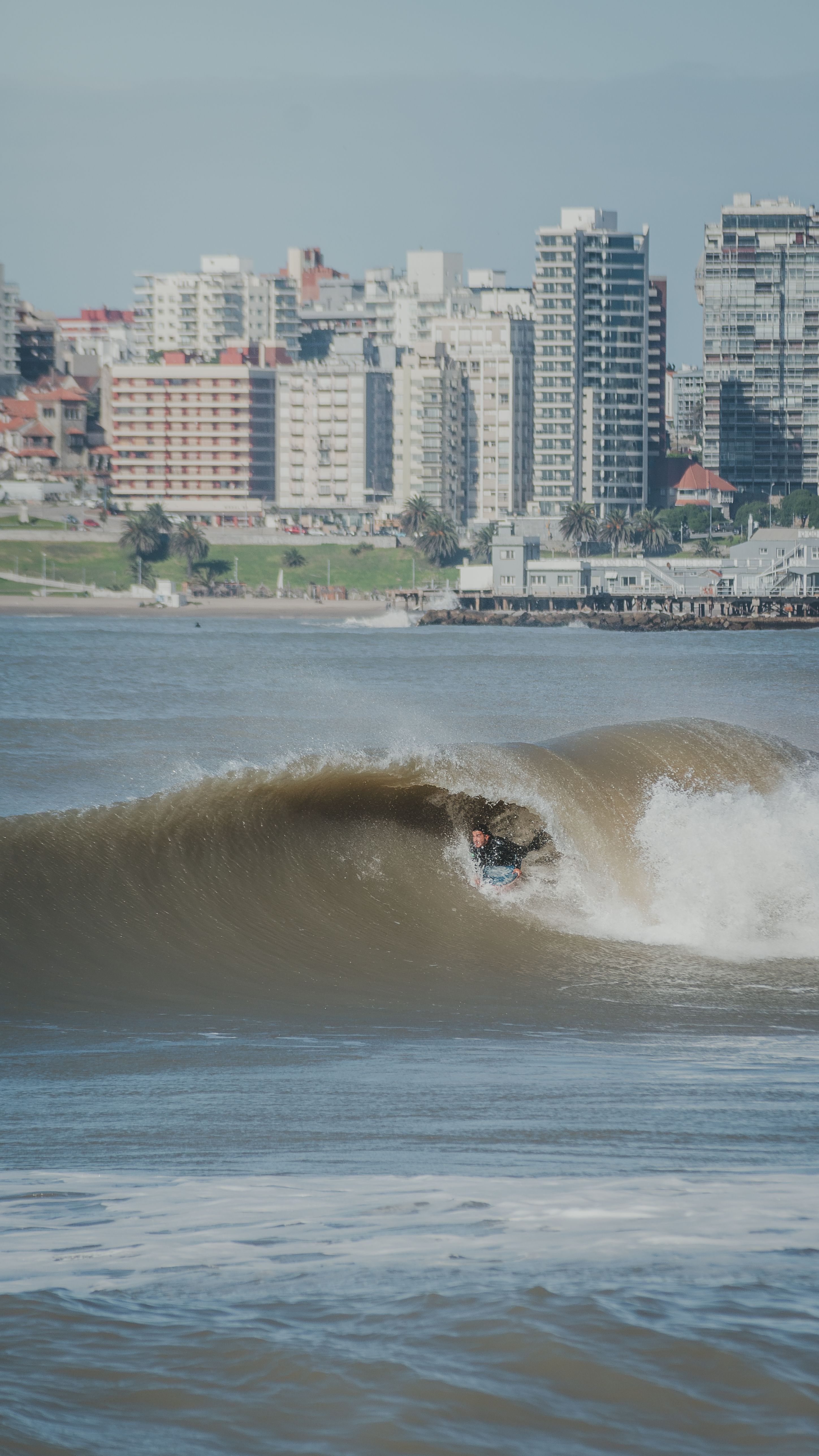 A surfer catches a wave near a bustling cityscape, blending nature with urban life.