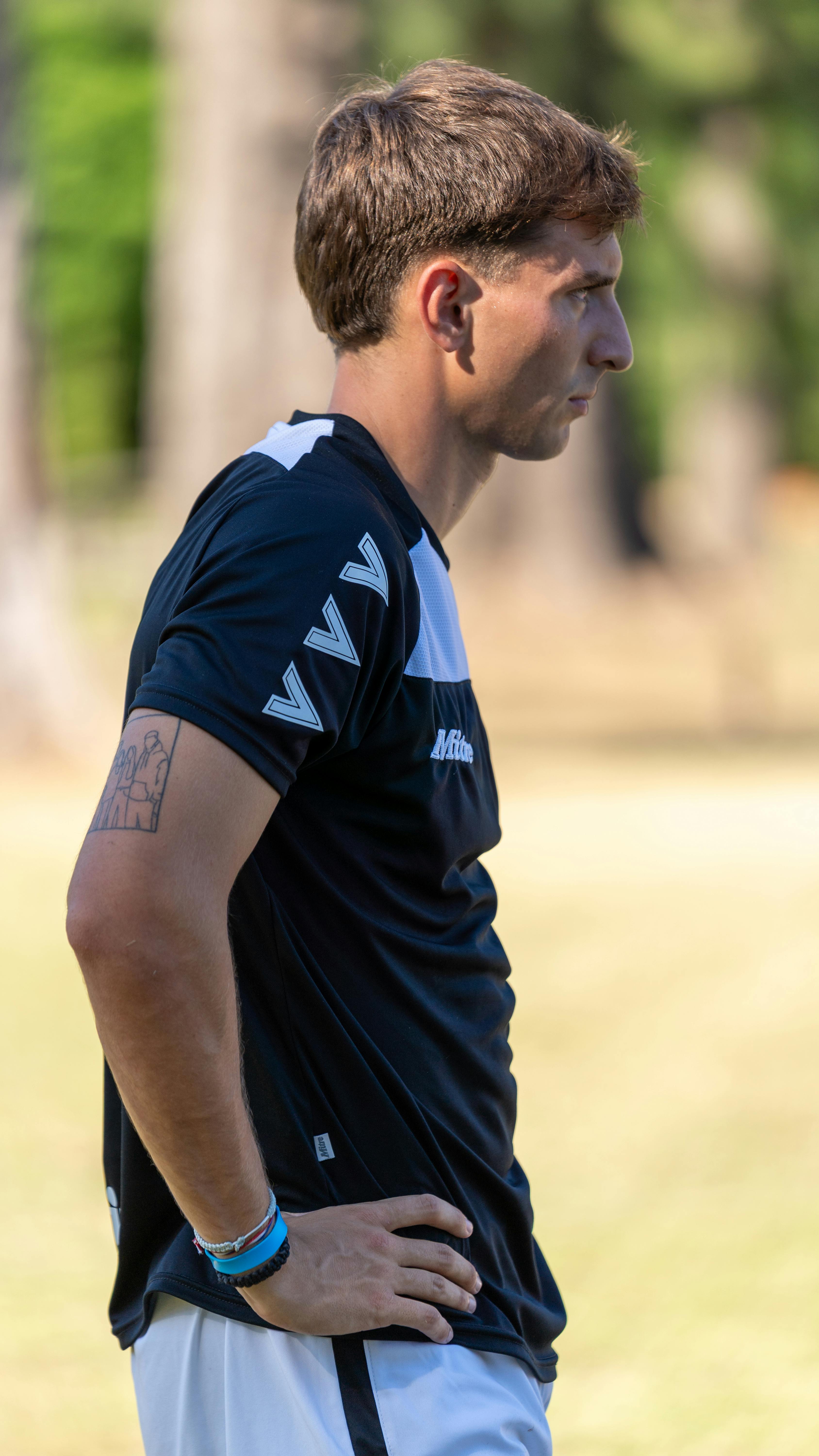 A man in sportswear standing outdoors in daylight, hands on hips, and facing right.