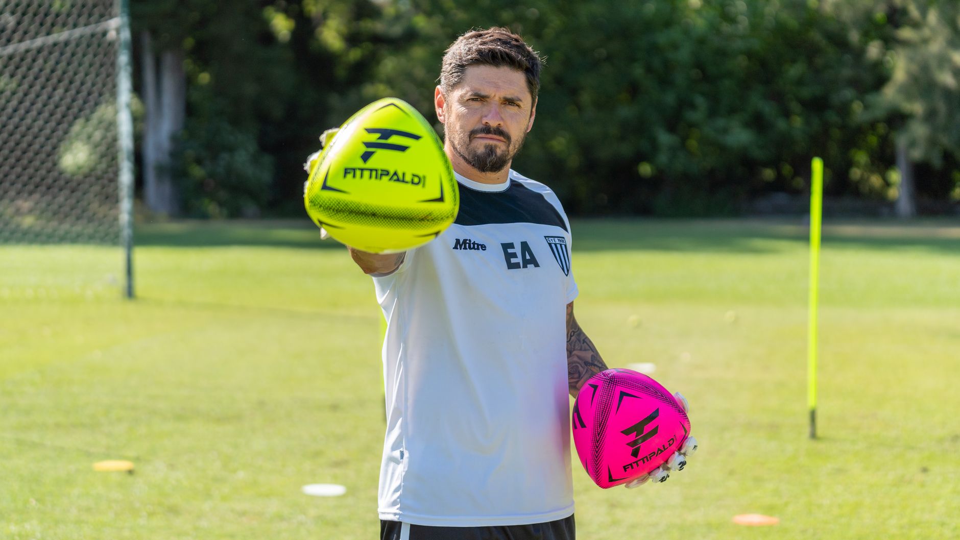Male coach on a sunny field holding neon yellow and pink footballs, ready for training.