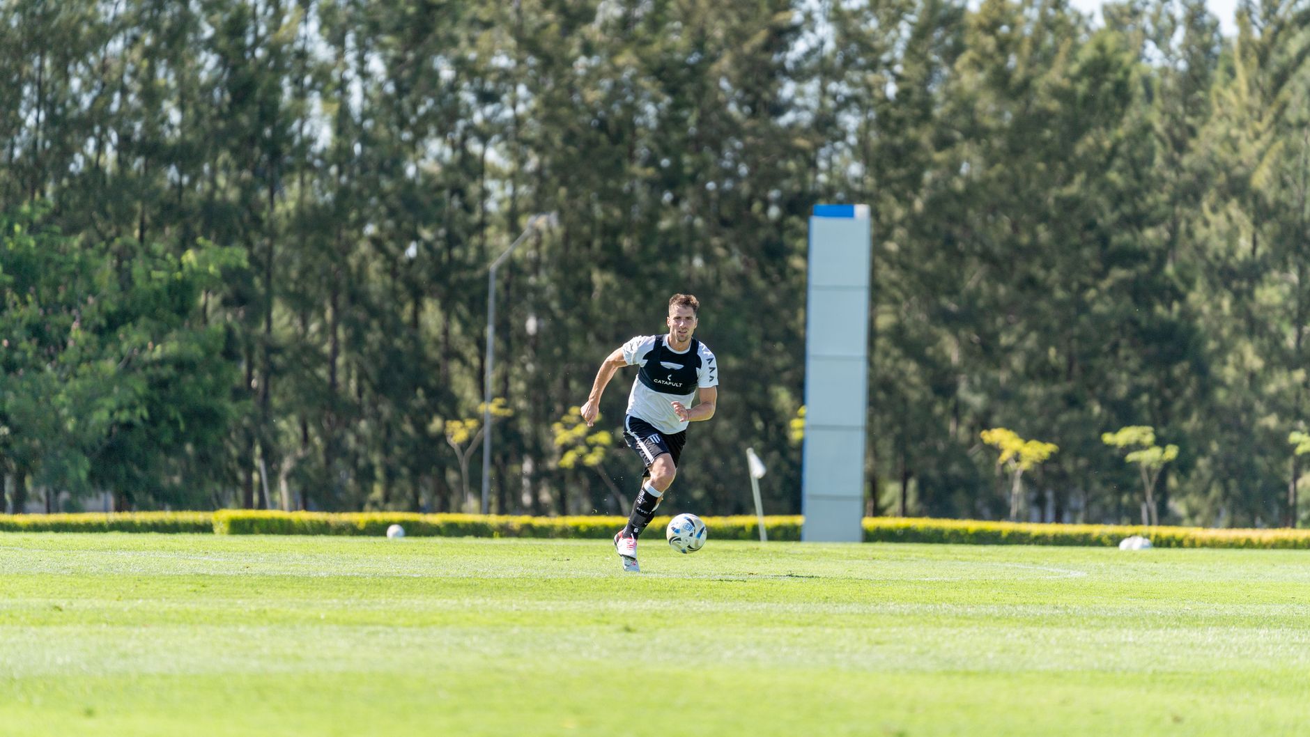 A soccer player in action dribbling a ball on a lush green field surrounded by trees on a sunny day.