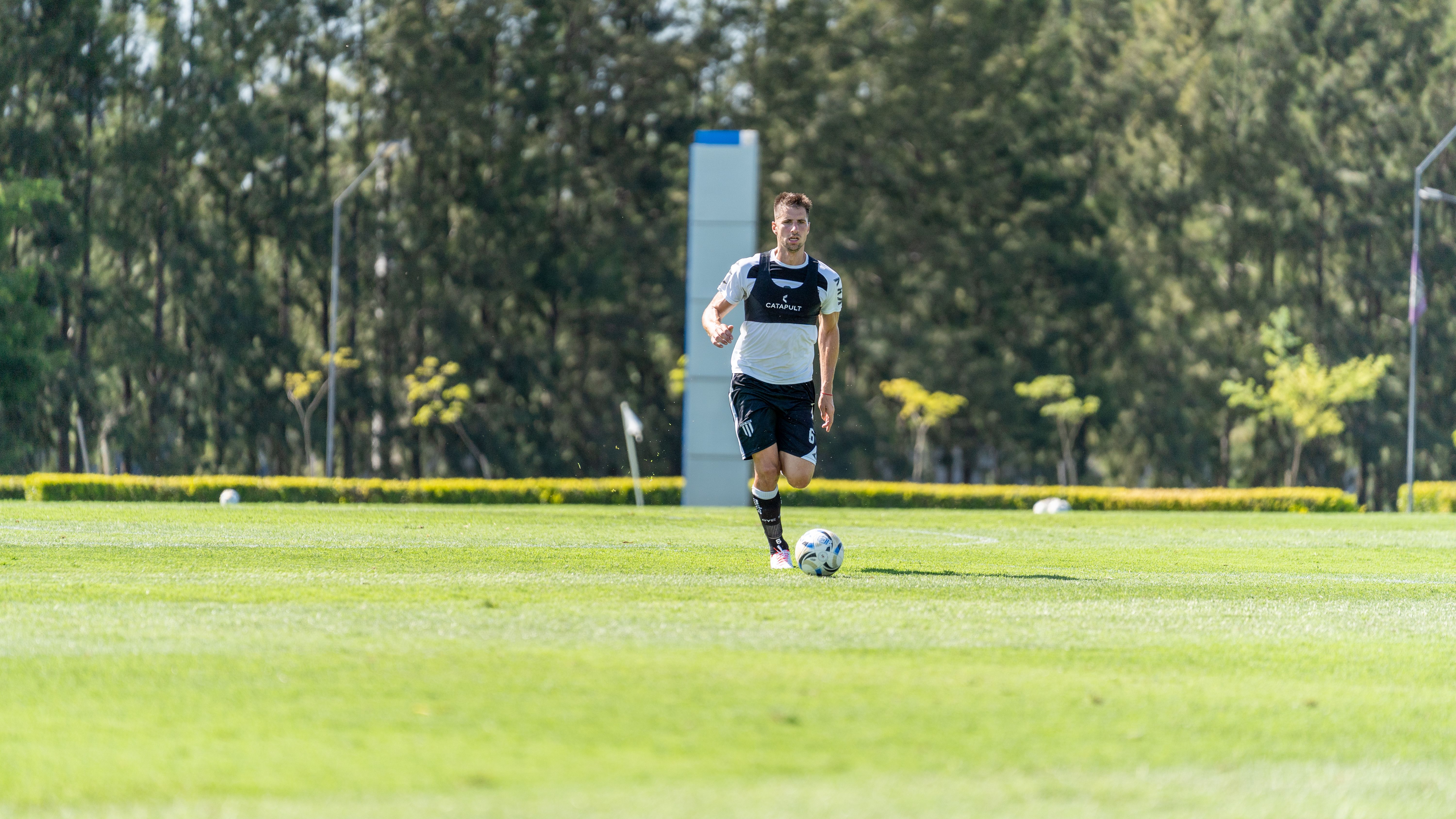 A soccer player in motion on a sunny day at a grassy field surrounded by trees.
