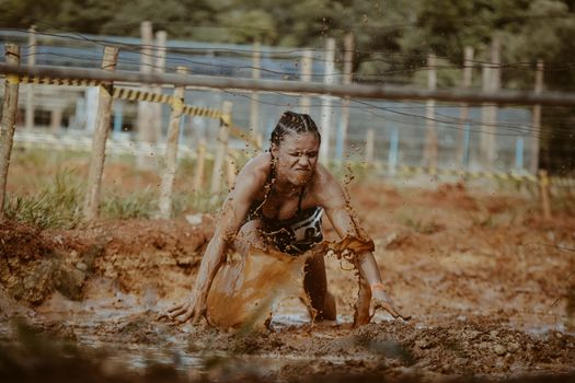 Determined woman crawls through muddy obstacle course in outdoor race challenge.
