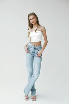 Elegant young woman in white top and jeans posing confidently in a minimalist studio setting.