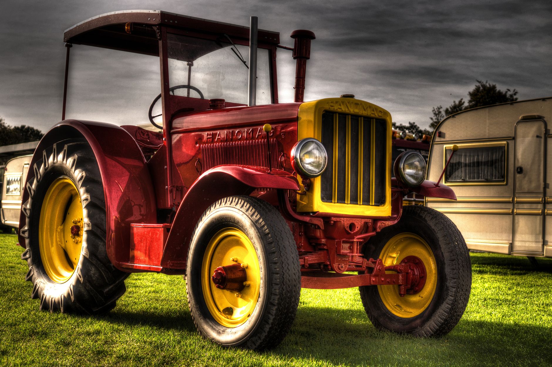 A vibrant red vintage tractor parked on a green field beside a caravan under a cloudy sky.