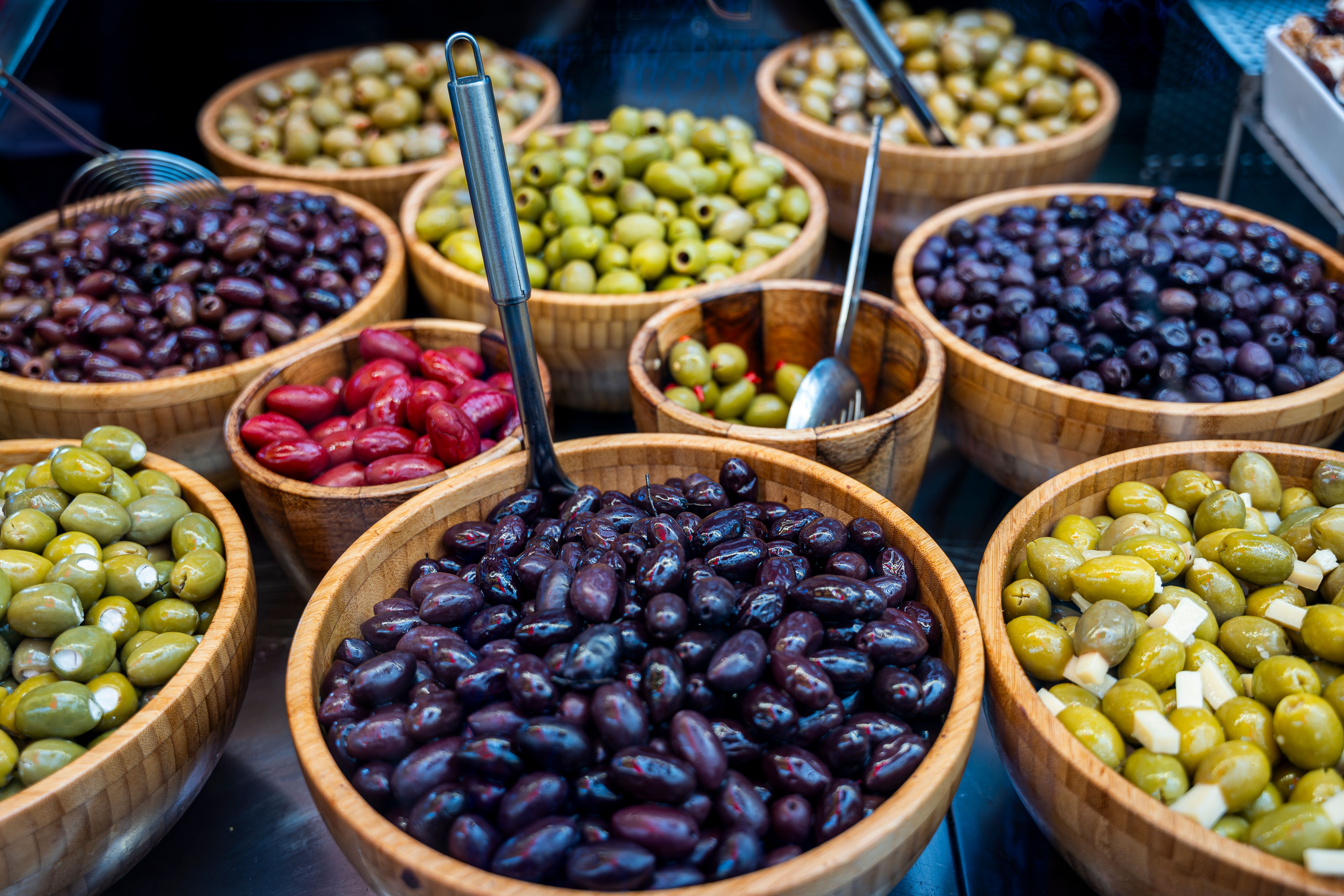 Various types of olives displayed in wooden bowls at an outdoor market.