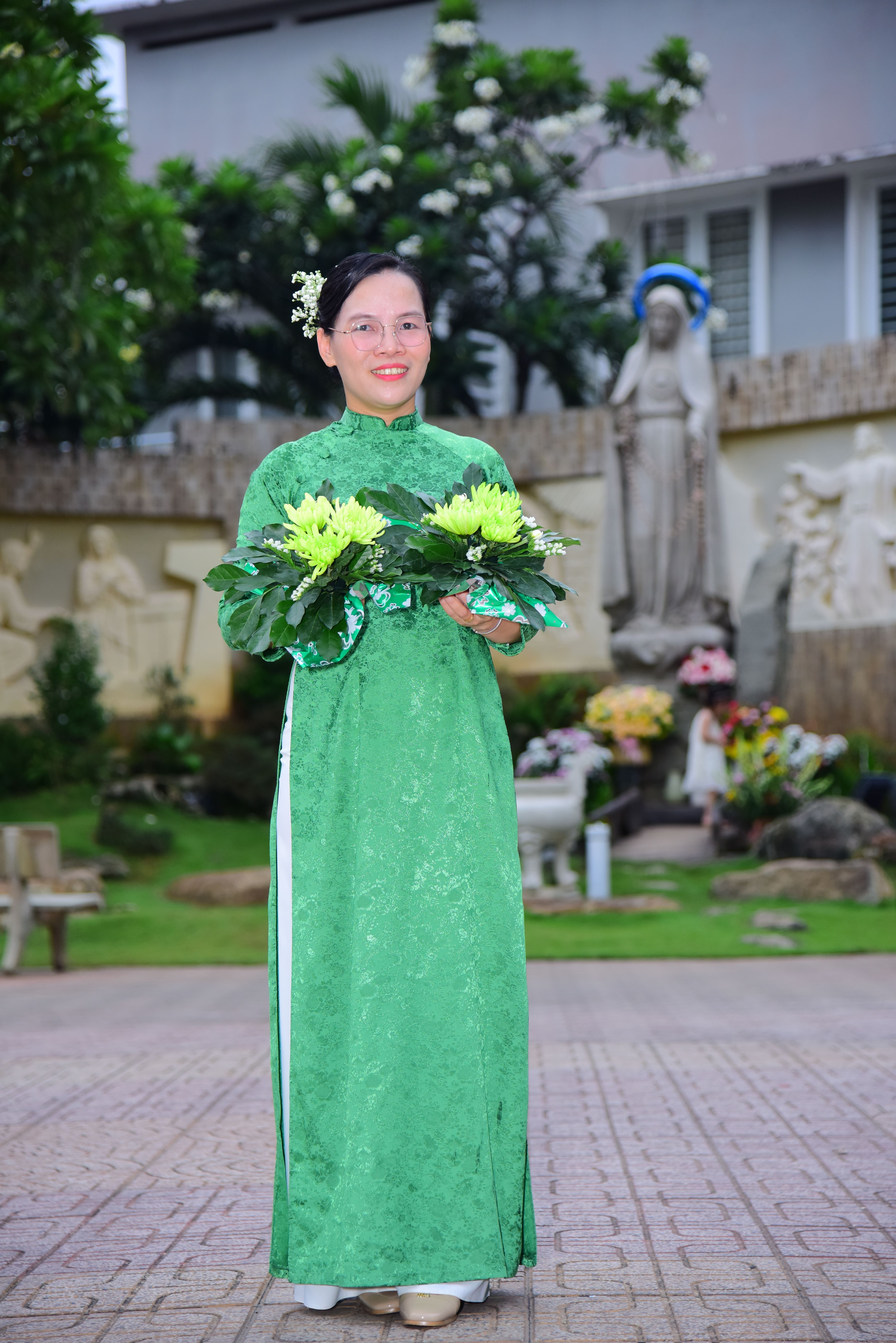 Woman in a vibrant green traditional dress holding a floral bouquet outdoors near a religious statue.