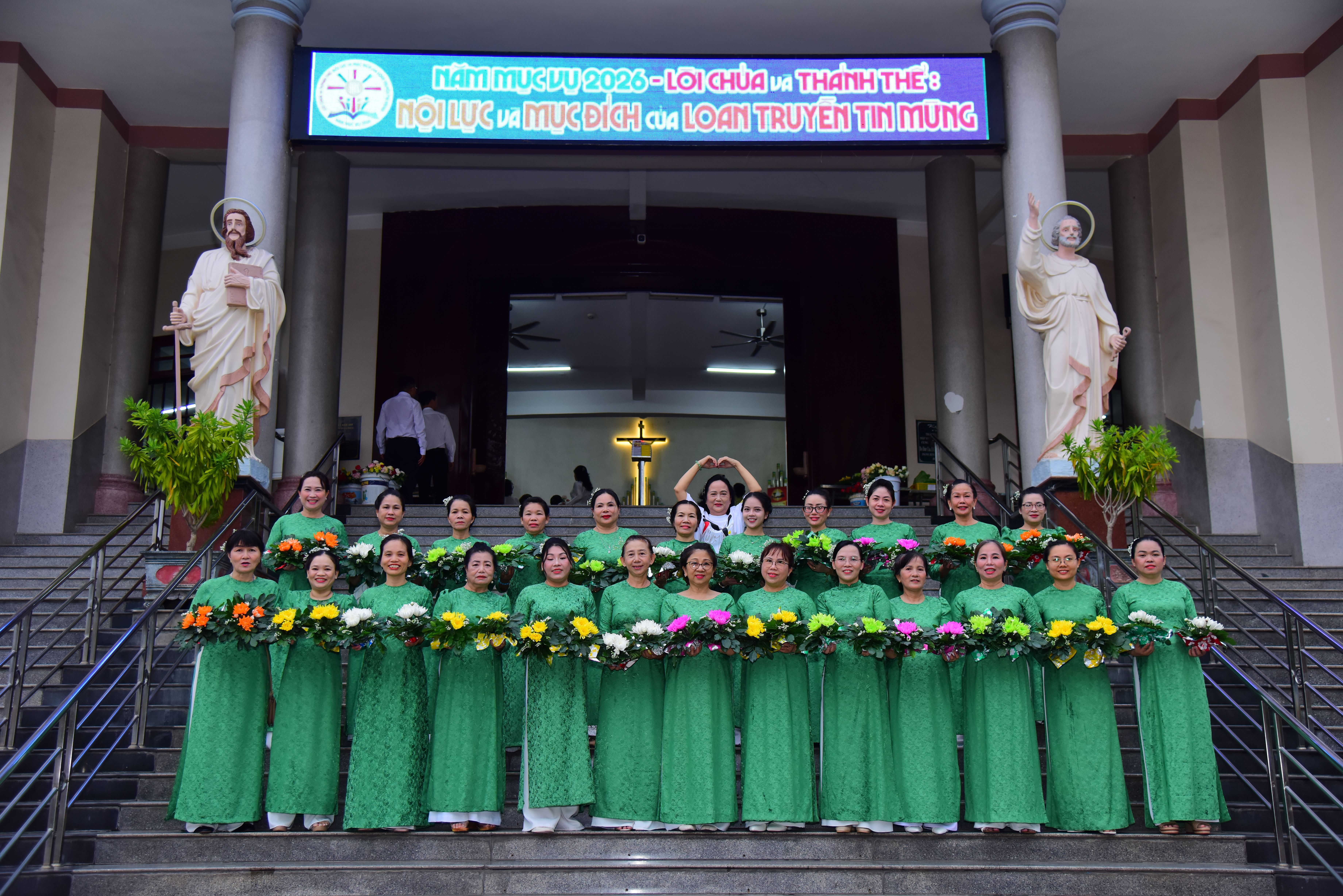 Women in green dresses pose with floral arrangements outside a church, sign above entrance.