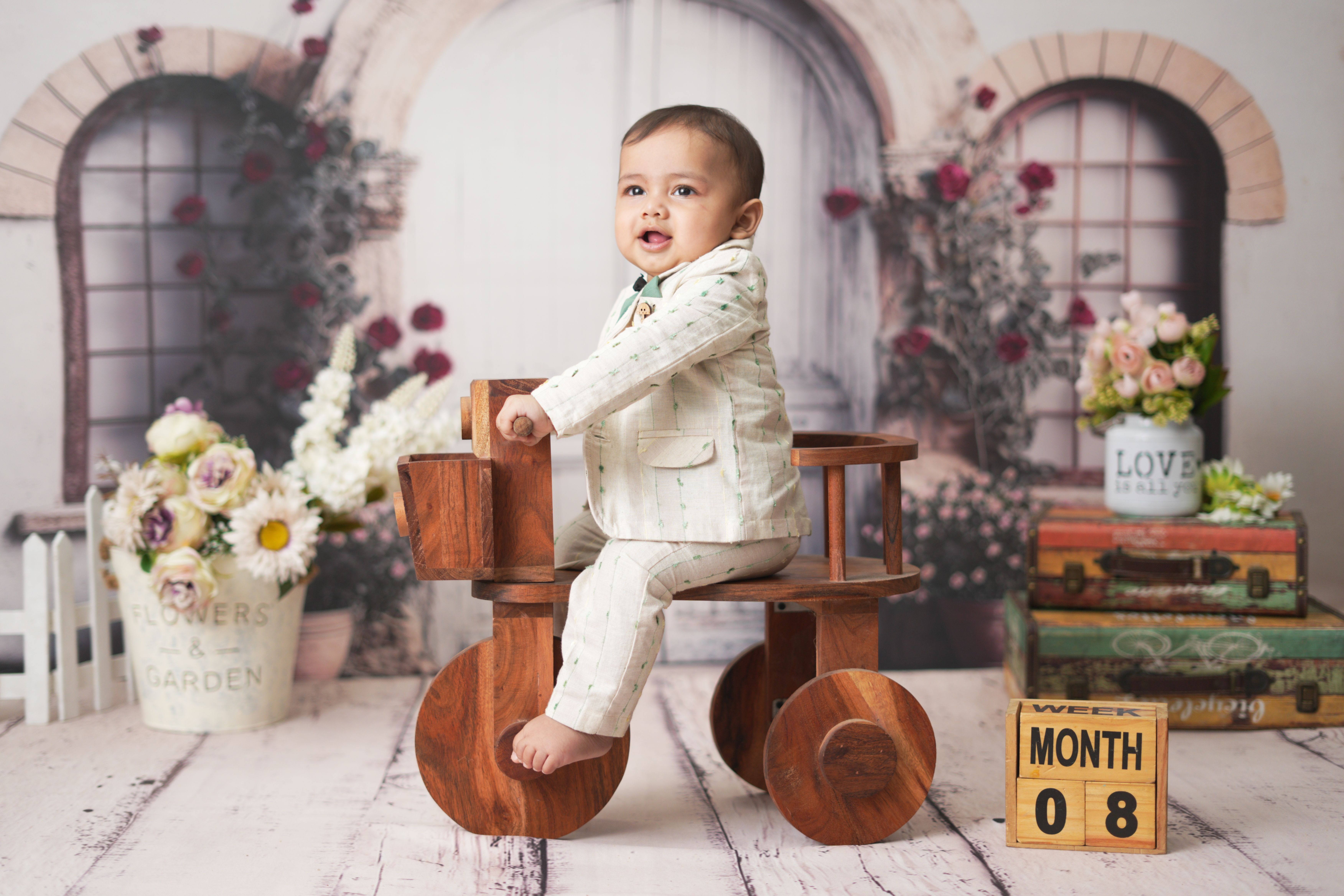 Adorable baby in stylish suit sitting on a wooden tricycle with floral background.