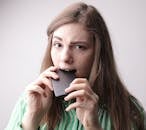 Nervous young female in casual clothing standing with cellphone in mouth after unpleasant phone call on gray background in studio