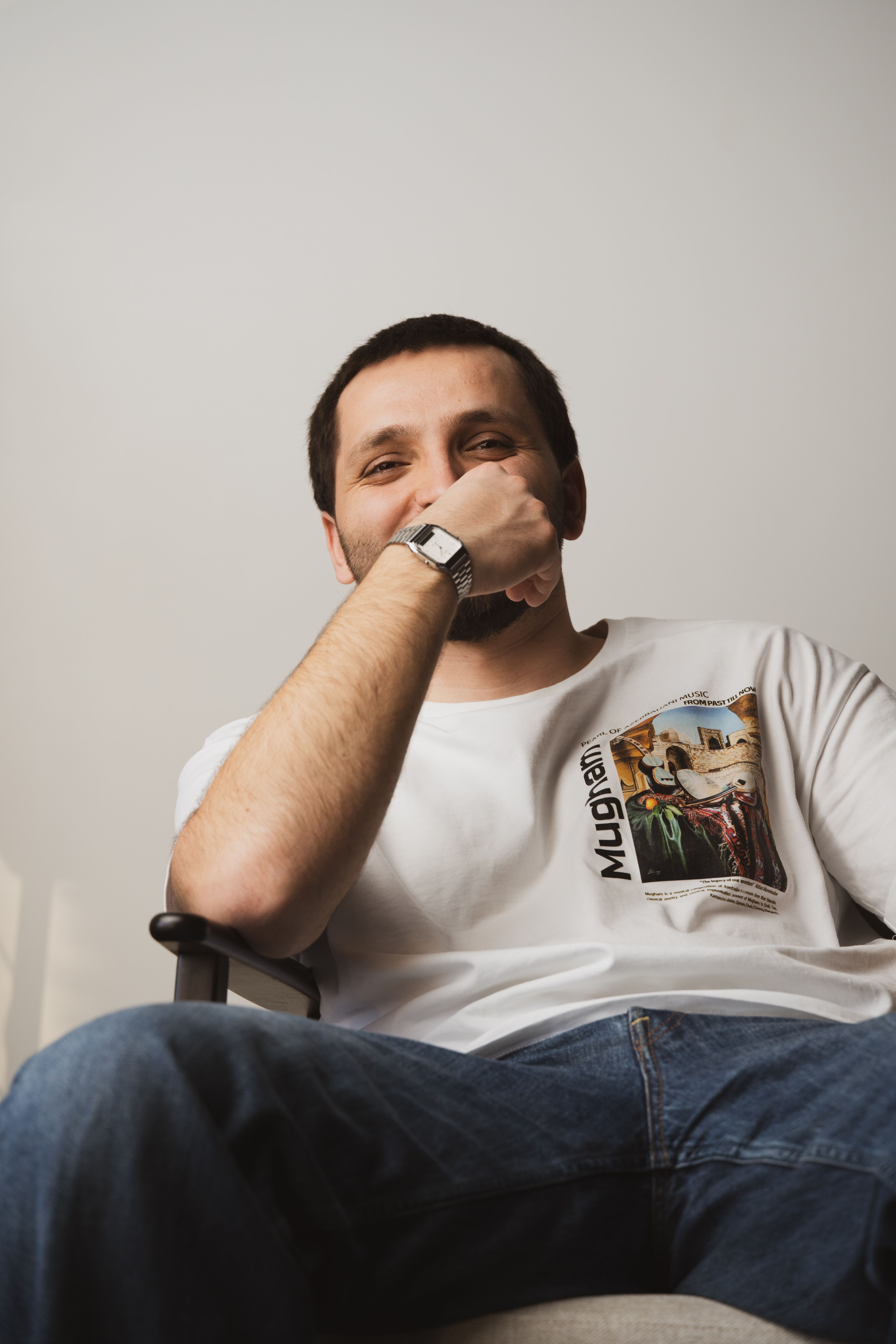 Young man sitting casually in a chair, wearing a printed T-shirt and jeans, with a relaxed expression.