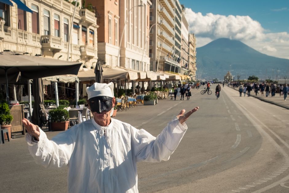 Pulcinella performer entertains on a bustling street in Naples, with Mount Vesuvius in the background.