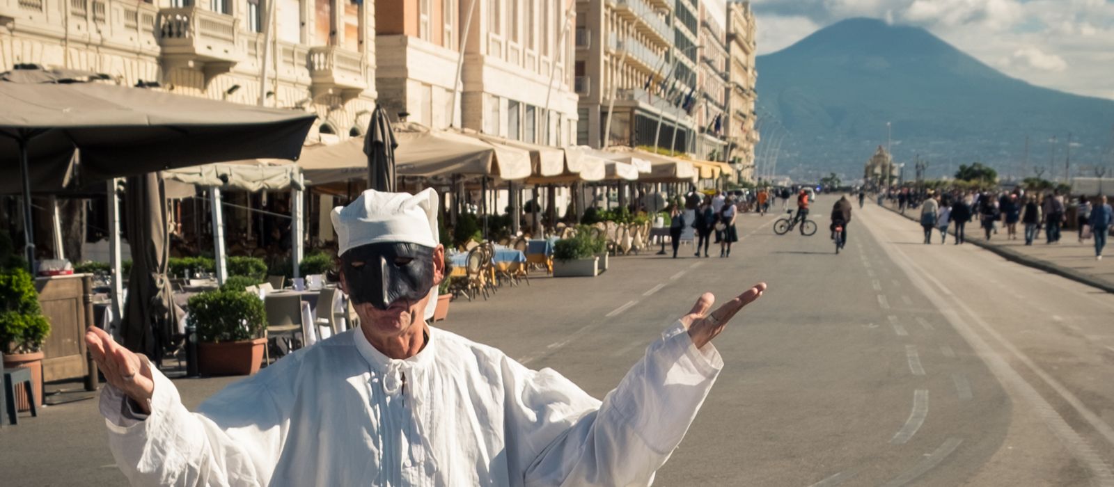 Pulcinella performer entertains on a bustling street in Naples, with Mount Vesuvius in the background.