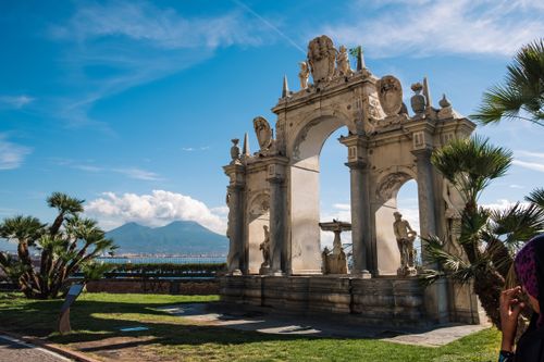 View of a historic arch in Naples with Mount Vesuvius and palm trees under a clear blue sky.