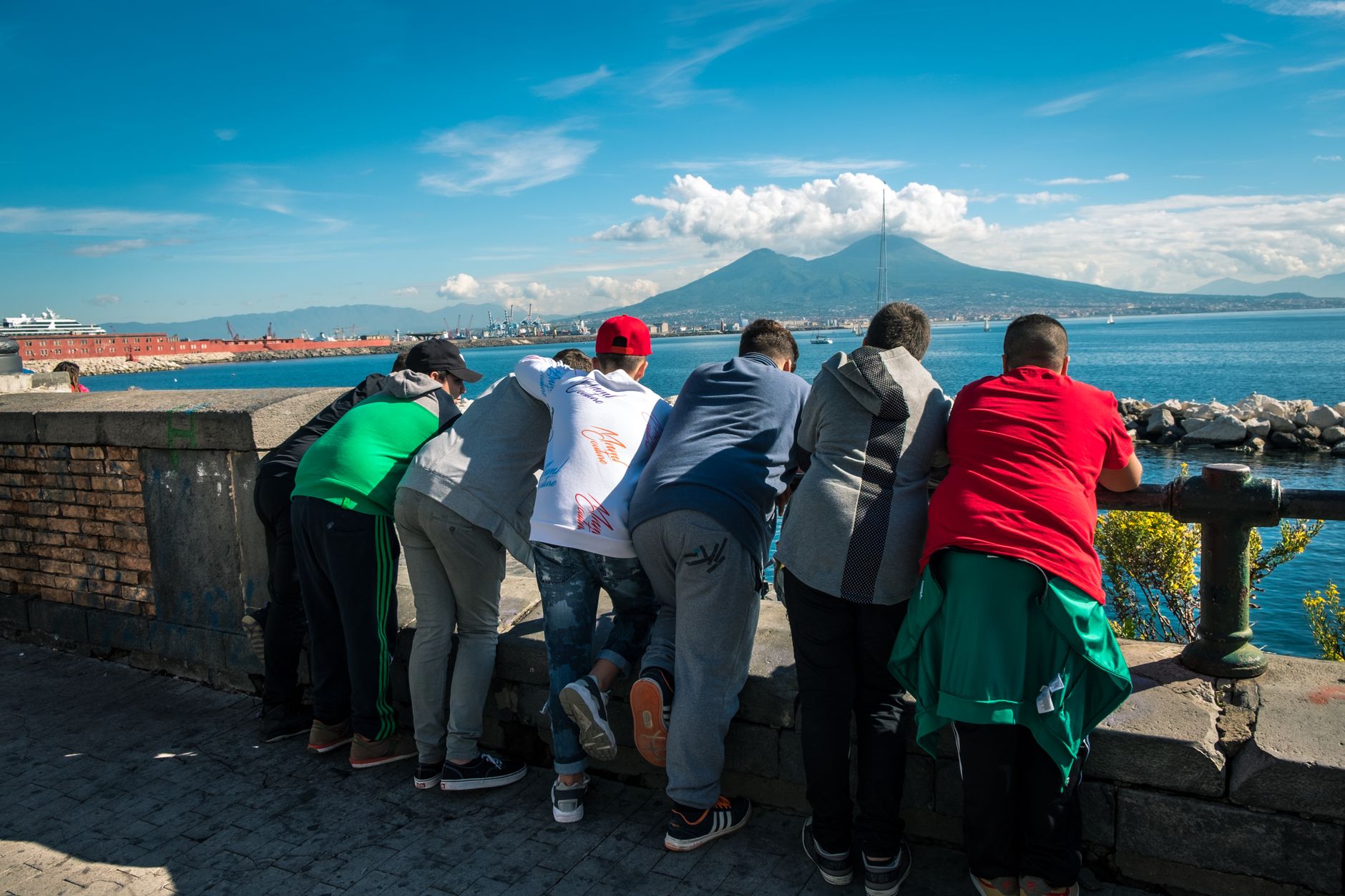 A group of young people looking towards Mount Vesuvius across the Naples waterfront on a sunny day.