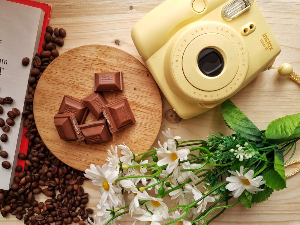 Free Top view of delicious pieces of milk chocolate bar with filling on wooden board near heap of aromatic coffee beans and instant camera with artificial chamomiles on table Stock Photo