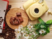 Top view of delicious pieces of milk chocolate bar with filling on wooden board near heap of aromatic coffee beans and instant camera with artificial chamomiles on table