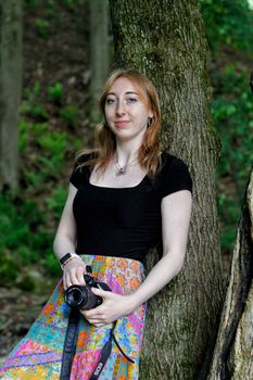 Woman holding a camera, leaning against a tree in a forest